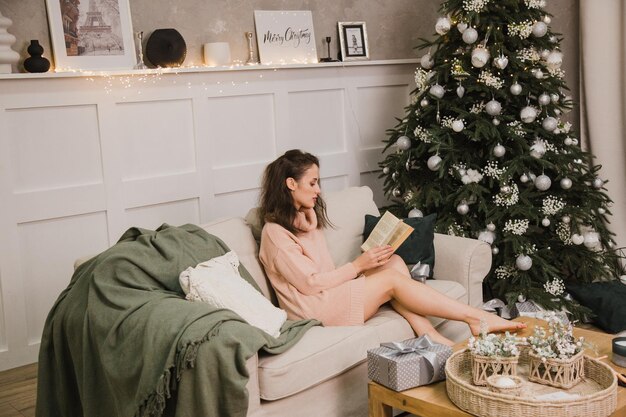 Een mooie vrouw zit op de bank bij de kerstboom en leest een boek. Nieuwjaar en Kerstmis