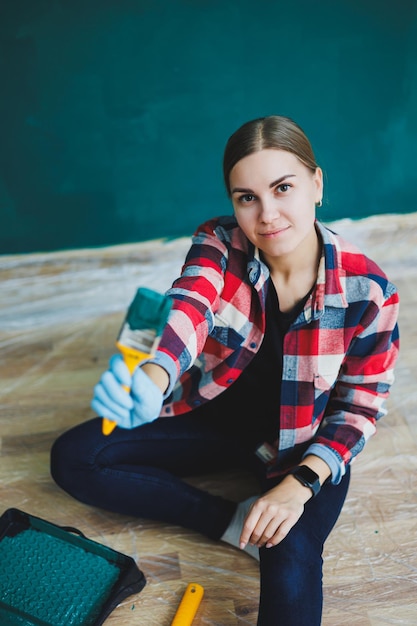 Een mooie vrouwelijke ontwerper maakt reparaties, schildert een grijze muur met groene verf met een penseel Geruit overhemd, lang haar en spijkerbroek Reparatie in het appartement