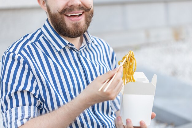 Een niet geïdentificeerde jonge mannelijke hipster met een snor en baard eet Chinese noedels met houten