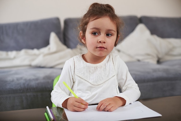 Een nieuwsgierig mooi klein meisje, zittend aan een tafel, houdt een pen vast en kijkt weg. Thuis tekenen.