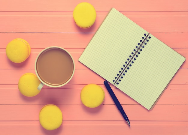 Een notebook met een pen, een kopje koffie en gele bitterkoekjes op een roze houten tafel.