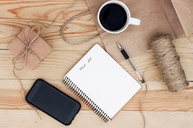 Een notitieboekje met een inscriptie Checklist op houten tafel. Het is omgeven door telefoon, zwarte pen, kopje koffie, cadeau, touw en een papieren zak.