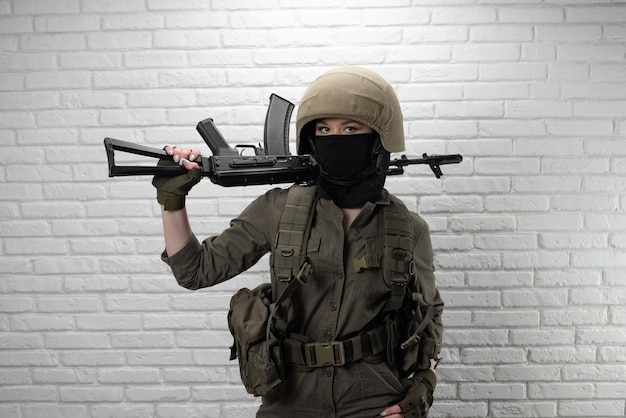 Een Oekraïense meisjessoldaat in een helm en militaire munitie met een Kalashnikov-aanvalsgeweer op de achtergrond van een bakstenen muur