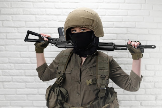Een Oekraïense meisjessoldaat in een helm en militaire munitie met een Kalashnikov-aanvalsgeweer op de achtergrond van een bakstenen muur
