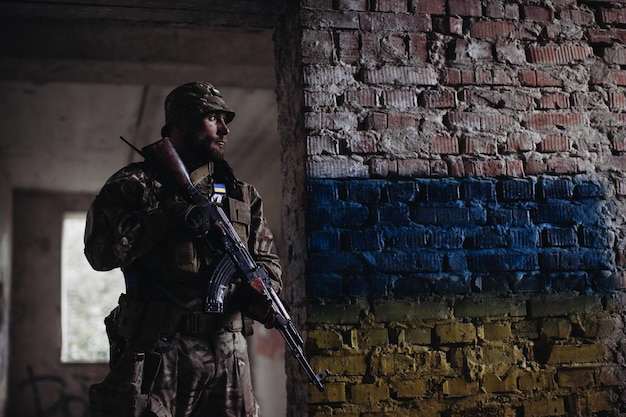 Een Oekraïense militaire man met een wapen in zijn handen staat tegen de achtergrond van de vlag van Oekraïne De vlag van Oekraïne is geschilderd op een bakstenen muur