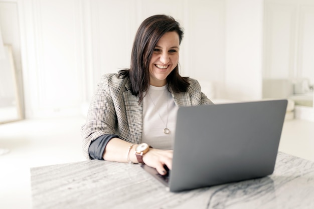Een online leraar gebruikt een laptop van een brunette vrouw met een stijlvolle bril en een jas die binnenshuis glimlacht