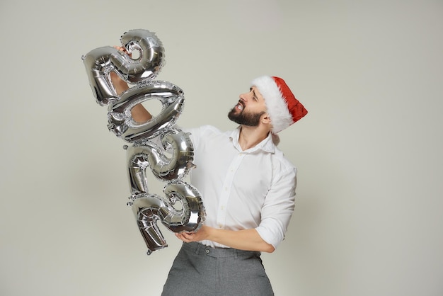 Een onstuimige man met een roodfluwelen kerstmuts duwt zilveren ballonnen in de vorm van 2022 naar links. Een man met een baard op een nieuwjaarsfeestje.