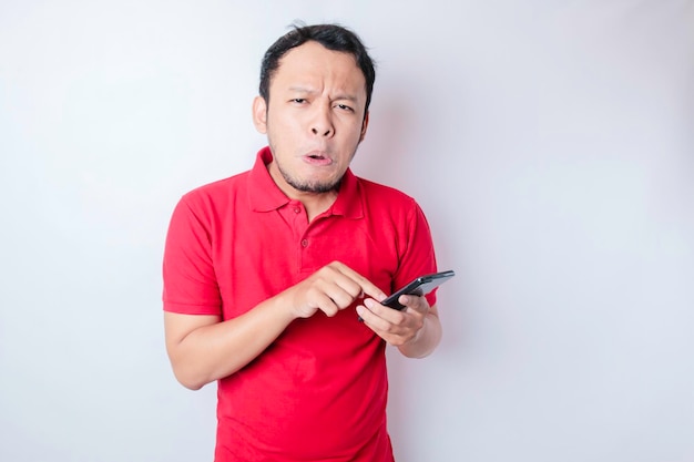 Een ontevreden jonge Aziatische man ziet er ontevreden uit met een rood t-shirt met geïrriteerde gezichtsuitdrukkingen die zijn telefoon vasthouden