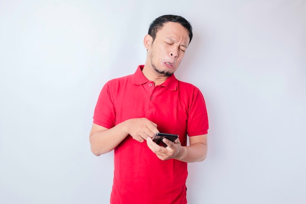 Een ontevreden jonge Aziatische man ziet er ontevreden uit met een rood t-shirt met geïrriteerde gezichtsuitdrukkingen die zijn telefoon vasthouden