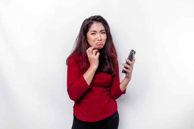 Een ontevreden jonge Aziatische vrouw ziet er ontevreden uit met rode, geïrriteerde gezichtsuitdrukkingen die haar telefoon vasthouden