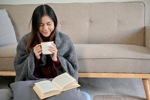 Een ontspannen Aziatische vrouw in een gezellige trui drinkt warme chocolade en leest een boek