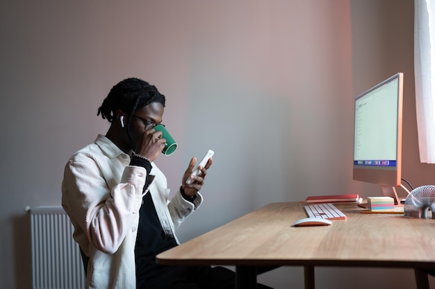 Een ontspannen, zorgeloze man die thee drinkt, zit aan een bureau en het gebruik van een mobiele telefoon zit aan tafel met een computer