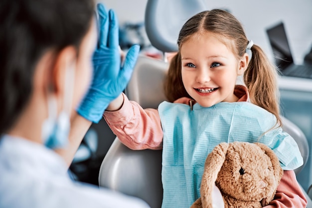 Foto een openhartige emotionele foto van een kind dat in een tandartsstoel zit, een speelgoedkonijn vasthoudt en vrolijk een highfive geeft aan de verpleegster. kindertandheelkunde