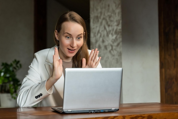 Een opgewonden zakenvrouw in een wit jasje viert succes op de werkplek door haar handen op te steken en naar een laptop te kijken