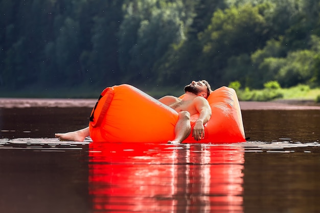 Een oranje opblaasbare ligstoel drijft langs de rivier, knappe bebaarde jongeman slaapt in een ligstoel, rustend op de natuur tijdens vakantie.