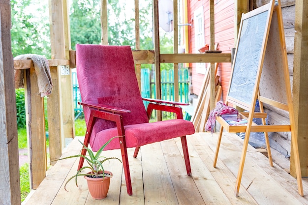 Een oude vintage fauteuil op een open veranda op een plankenvloer, naast een ezel en een aloë bloempot.