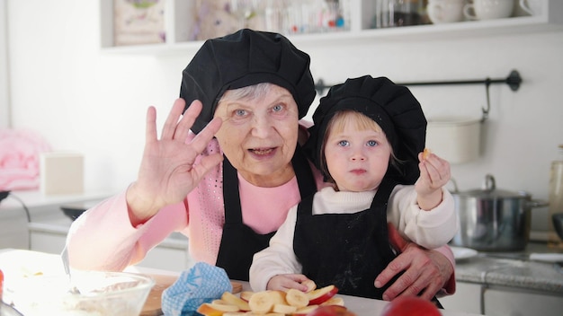 Een oude vrouw met een klein meisje dat met haar handen zwaait en in de keuken in de camera kijkt