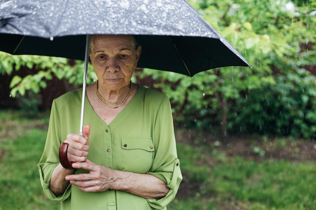 Een oude vrouw staat op straat onder de regen met een zwarte paraplu