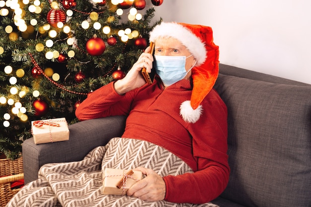 Een oudere blanke man in een kerstmuts met een medisch masker spreekt aan de telefoon, zittend op een bank in de buurt van een kerstboom. Zelfisolatie, coronavirus, vakantiestemming. Geschenkdozen.