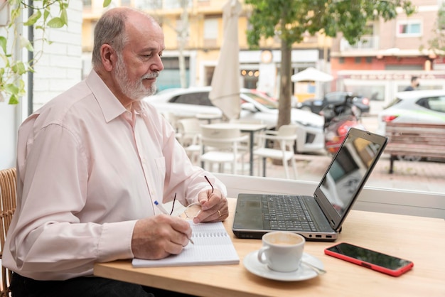 Een oudere man die aan een tafel zit met zijn laptop ervoor, de gegevens controleert en aantekeningen maakt in zijn notitieboekje, koffiekopje en mobiele telefoon naast hem