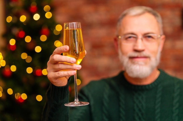 Een oudere man in een groene gebreide trui die een toast uitbrengt met champagne bij de kerstboom