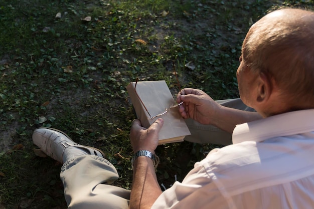 Een oudere man in een wit overhemd zit op een deken op de grond in een park en leest een interessant boek Een gepensioneerde alleen rust in de natuur gepassioneerd over zijn hobby