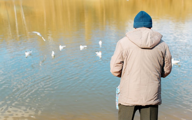 Een oudere man in jas en hoed die op de kust in de buurt van water staat en vogels buiten voedt Achteraanzicht kopieerruimte