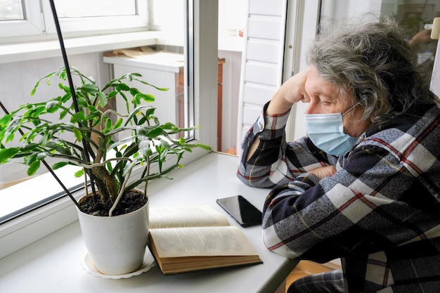 Een oudere man met een beschermend masker op zelfisolatie die thuis een boek leest terwijl hij bij het raam zit Gezondheidszorgconcept