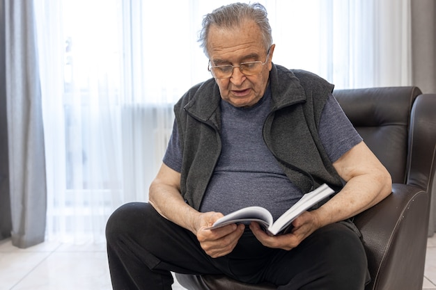 Een oudere man met een bril leest een boek terwijl hij op een fauteuil zit.