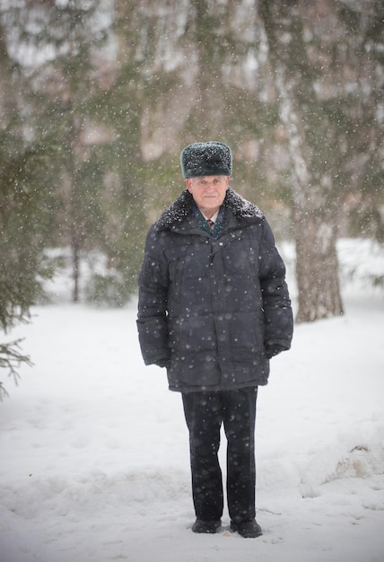 Een oudere man met een warme muts die buiten in het winterbos staat terwijl sneeuwval een donsjack draagt