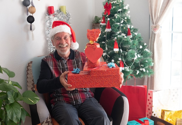 Een oudere man met een witte baard en een kerstmuts die lacht van geluk en veel kerstcadeautjes vasthoudt.