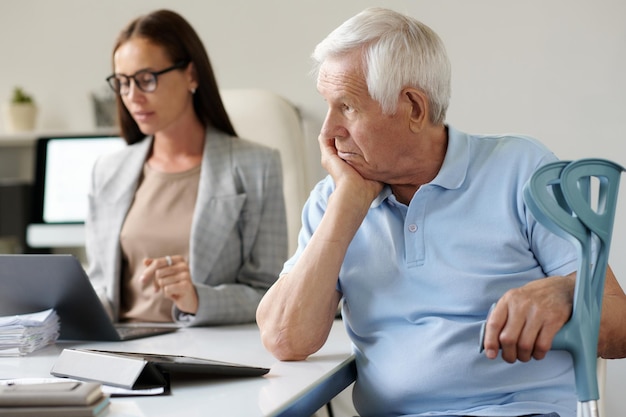 Foto een oudere man met krukken zit aan tafel met een maatschappelijk werker in het kantoor.