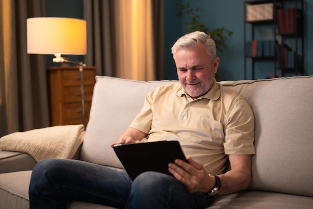 Een oudere man speelt op een tablet terwijl hij 's avonds thuis op de bank in de woonkamer zit man