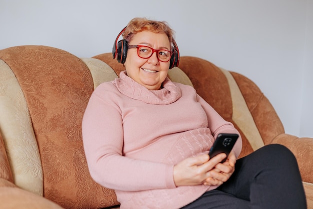 Foto een oudere vrouw die comfortabel op haar bank zit en naar haar favoriete nummers luistert met haar koptelefoon en er gelukkig en ontspannen uitziet thuis glimlachende oudere dame ontspannen op de bank met koptelefoon aan