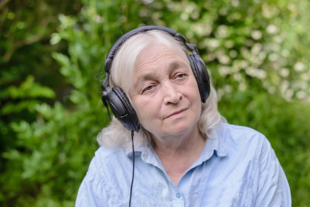 Een oudere vrouw in een blauw shirt die naar muziek luistert op een koptelefoon in de tuin, in de natuur