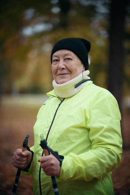 Een oudere vrouw in een groen donsjack staat in een park met stokken voor nordic walking