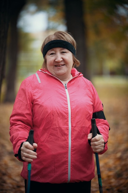 Een oudere vrouw in een roze donsjack staat in een park met stokken voor nordic walking