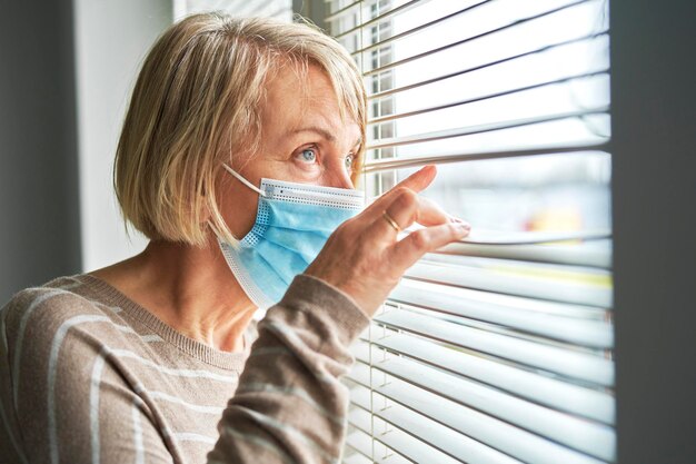 Een oudere vrouw in quarantaine die een masker draagt kijkt door de blinds.