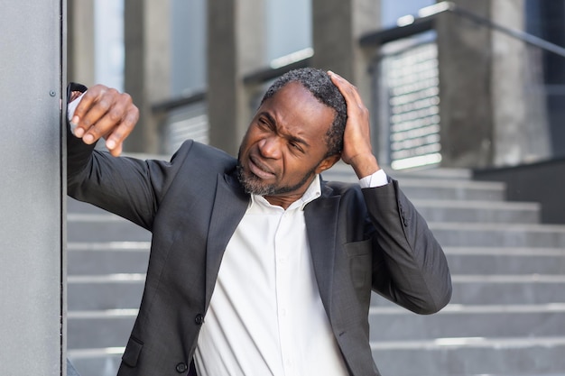 Een overstuur senior Afro-Amerikaanse man in een pak staat buiten een kantoorcentrum leunend tegen de muur