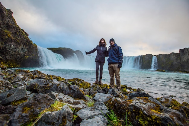 Een paar aan de onderkant van de Godafoss-waterval met zonsondergang op de achtergrond, IJsland