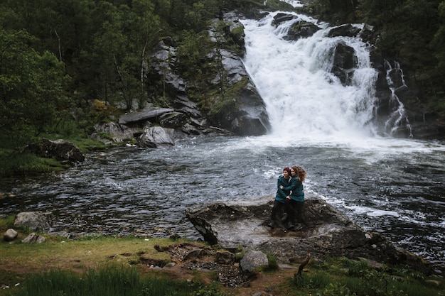 Een paar geliefden in groene regenjassen, zittend op een rots, tegen de achtergrond van een waterval