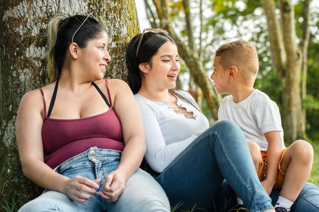 Een paar jonge latijns-vrouwen praten met hun jonge zoon die naast een boom in een park zit