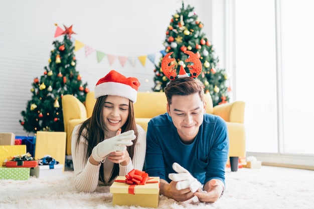 Een paar kerstcadeau concept Jonge gelukkige Aziatische vrouw met een kerstman hoed verrast haar vriendje met handschoenen terwijl ze op het tapijt ligt met een kerstboom op de achtergrond