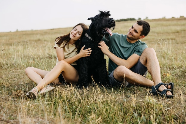 Een paar man en vrouw zitten buiten in een veld met een zwarte reusachtige schnauzer raszuivere hond die omhelst