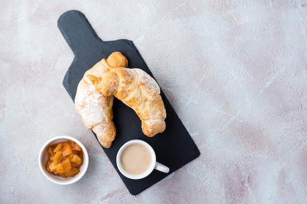 Een paar verse knapperige croissants op een leisteen, een kopje koffie en jam in een kom op een lichte tafel. Koffiepauze of ontbijt. Bovenaanzicht. Ruimte kopiëren