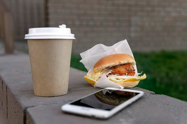 Een papieren beker met koffie of thee, een broodje kip en een smartphone liggen op een bankje op straat. Het concept van junkfood tijdens de lunchpauze