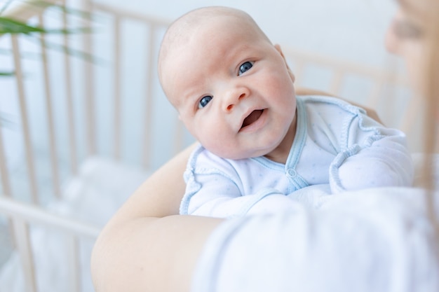 Een pasgeboren babyjongen in de armen van zijn moeder lacht bij de wieg thuis, ouderlijke liefde en zorg voor de baby in de eerste dagen na de geboorte.