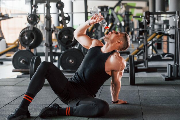 Een pauze nemen en water drinken Jonge sportieve sterke man in zwarte kleding heeft een trainingsdag in de sportschool