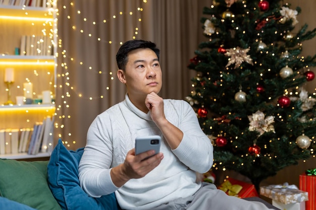 Een peinzende en serieuze jonge aziatische man zit thuis op de bank bij de kerstboom met behulp van a