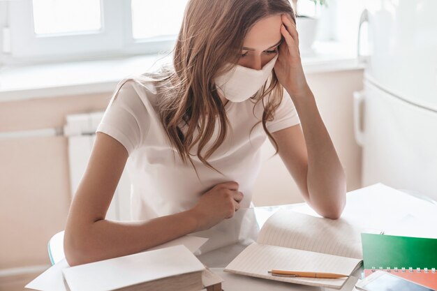 Een peinzende jonge vrouw met een medisch masker en een wit T-shirt zit thuis in de kamer aan tafel en werkt op afstand. Studentenmeisje die thuis, quarantaineisolatie bestuderen, het concept van het verblijfhuis